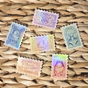 May include: A collection of six holographic stickers in a postage stamp design. Each sticker features a unique illustration and text, including the words "NOMAD," "HEARTHIAN," "ANGLERFISH," "OWLK," "PROTO," and "INSECTOID."