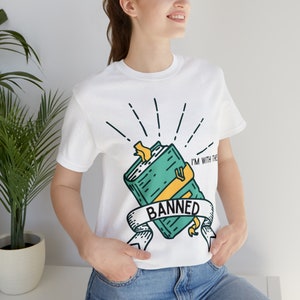 Pode incluir: Camiseta branca com uma ilustração de um livro verde e o texto "I'm with the Banned".