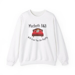 Könnte beinhalten: Weißer Pullover mit einem Aufdruck eines Bettes mit einer Krone und dem Text "Macbeth B&B" und "We'll Treat You Like Royalty".