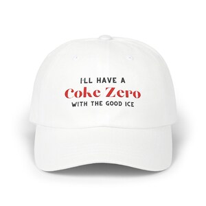 以下が含まれることがあります： 黒い文字で「I'll have a Coke Zero with the good ice」と書かれた白い野球帽。 「Coke Zero」という単語は赤色です。