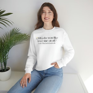 Puede incluir: Sudadera blanca de manga larga con el texto "OMG do you like love me at all?" en negro y amarillo, con "-Lady Macbeth Basically" debajo. La sudadera se combina con unos vaqueros azules.