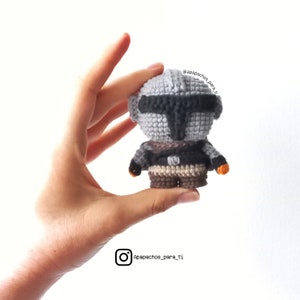 Puede incluir: Un personaje mandaloriano de crochet gris y marrón con una visera naranja. El personaje sostiene un pequeño objeto naranja en cada mano. El personaje está siendo sostenido en la mano de una persona.