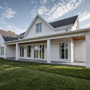 May include: Exterior view of a large, white house with a dark gray roof. The house has multiple windows and a covered porch with white columns. The lawn is green and well-manicured, with trees in the background. The sky is blue with scattered clouds.