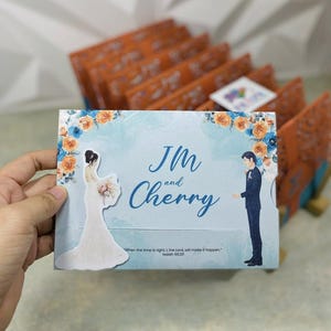 May include: A wedding invitation card featuring a bride and groom illustration, with the names "JM and Cherry" in elegant script. The card has a light blue background with floral accents and a quote from Isaiah 60:20.