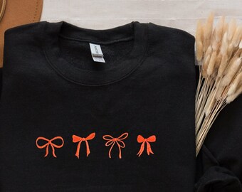 Puede incluir: Sudadera negra con cinco lazos rojos bordados. La sudadera tiene cuello redondo y mangas largas. El texto "EMBROIDERED" está en la parte superior de la imagen y "BLACK" en la parte inferior.