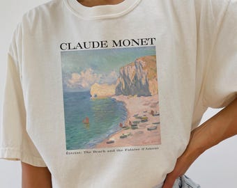 Op de afbeelding: Een wit T-shirt met een grafische print van een schilderij van Claude Monet. Het schilderij toont een strandscene met een klif op de achtergrond. De tekst "Etretat: The Beach and the Palaise d'Amont" is onder het schilderij gedrukt.