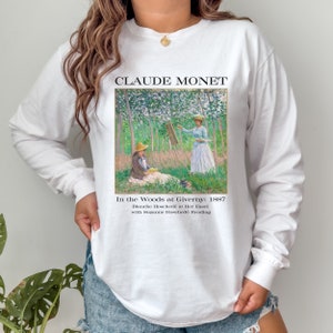 May include: White long-sleeve shirt with a graphic print of Claude Monet's painting "In the Woods at Giverny: 1887". The text "Blanche Hosched&eacute; at Her Easel with Suzanne Hosched&eacute; Reading" is printed below the painting.