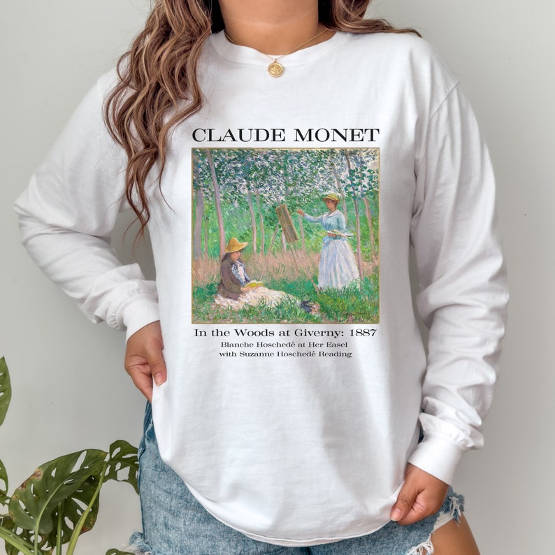 May include: White long-sleeve shirt with a graphic print of Claude Monet's painting "In the Woods at Giverny: 1887". The text "Blanche Hosched&eacute; at Her Easel with Suzanne Hosched&eacute; Reading" is printed below the painting.