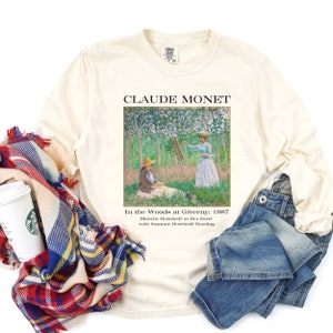May include: A beige long-sleeve t-shirt with a print of Claude Monet's painting "In the Woods at Giverny: 1887". The text "Claude Monet" is printed above the painting and the text "In the Woods at Giverny: 1887 Blanche Hosched&eacute; at Her Easel with Suzanne Hosched&eacute; Reading" is printed below the painting.