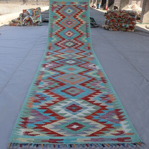 May include: A long, narrow rug with a geometric pattern in shades of blue, red, green, and white. The rug has a fringe on both ends.