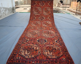 May include: An antique runner rug with a complex geometric design in red, orange, and navy blue. The rug's pattern includes repeating square and diamond shapes, creating a traditional, ornate look. The rug is laid out on a light blue sheet.