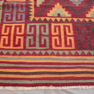 May include: A close-up of a colorful rug with geometric patterns. The rug features a red background with orange, green, and brown geometric shapes. The border has a fringe. The rug is likely made of woven fabric.
