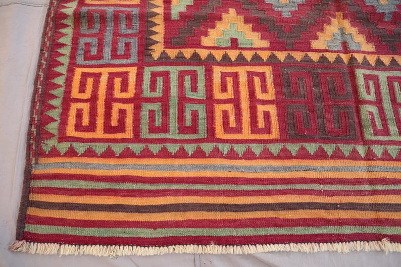May include: A close-up of a colorful rug with geometric patterns. The rug features a red background with orange, green, and brown geometric shapes. The border has a fringe. The rug is likely made of woven fabric.