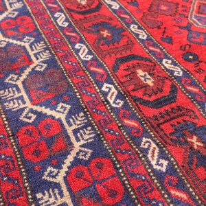 May include: Close-up of a vibrant red rug with intricate geometric patterns. The rug features a central field of red, accented by navy blue and beige borders with repeating designs. The detailed craftsmanship is evident in the woven texture.