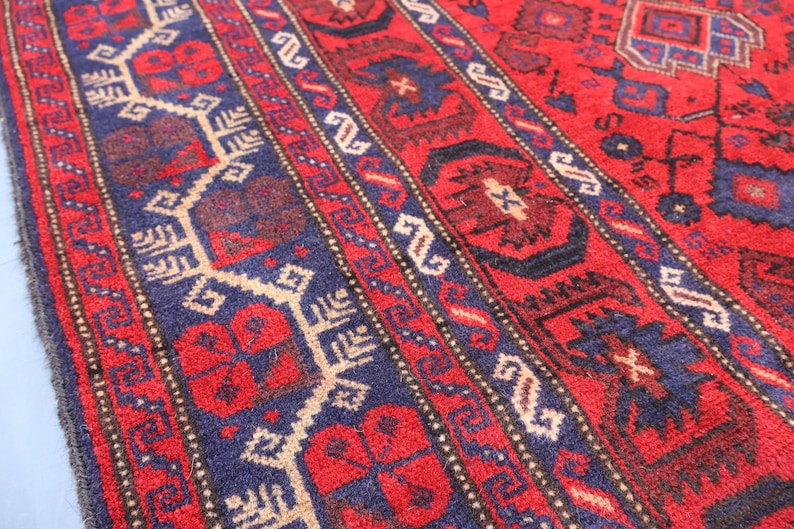 May include: Close-up of a vibrant red rug with intricate geometric patterns. The rug features a central field of red, accented by navy blue and beige borders with repeating designs. The detailed craftsmanship is evident in the woven texture.