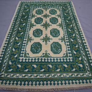 Puede incluir: Alfombra rectangular beige con un patrón geométrico verde y turquesa. La alfombra presenta un diseño central de octágonos y cruces, enmarcado por un borde de hojas estilizadas y formas de diamante. La alfombra tiene una franja en el borde inferior.