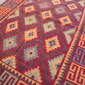 May include: A rectangular rug with a geometric pattern. The rug features a deep red background with repeating diamond shapes in orange, teal, and gray. The border has a repeating geometric design in orange and red.