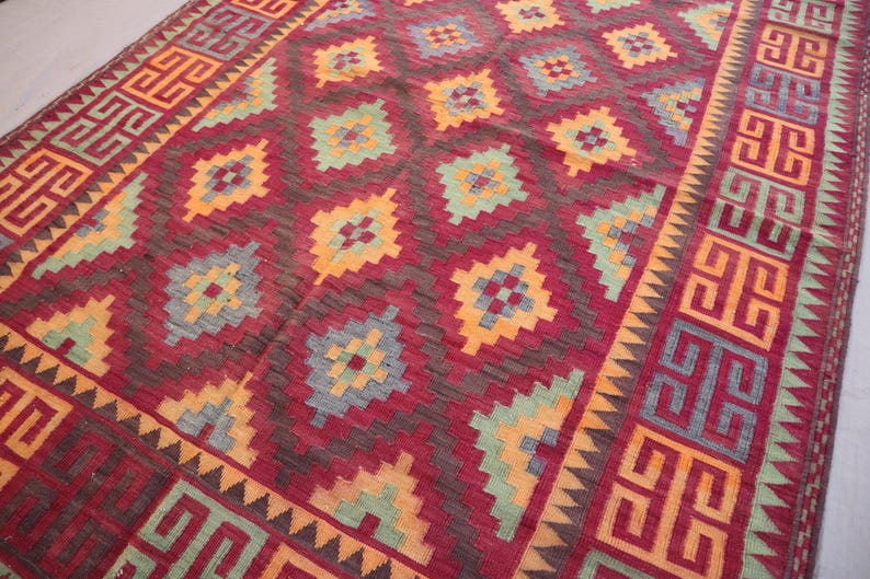 May include: A rectangular rug with a geometric pattern. The rug features a deep red background with repeating diamond shapes in orange, teal, and gray. The border has a repeating geometric design in orange and red.