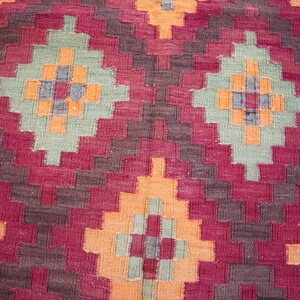 May include: Close-up of a woven rug with a geometric pattern. The rug features a repeating design of diamond shapes in shades of red, orange, green, and gray. The texture of the rug is visible, suggesting a handmade or artisanal quality.