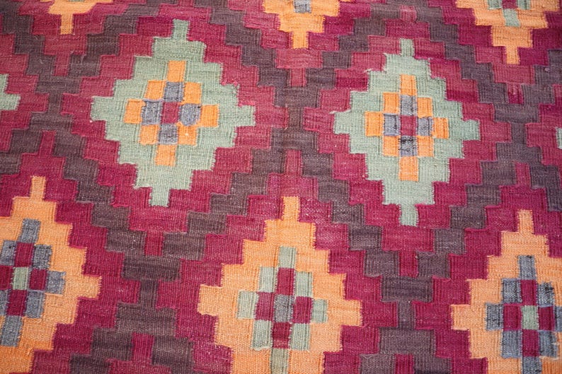 May include: Close-up of a woven rug with a geometric pattern. The rug features a repeating design of diamond shapes in shades of red, orange, green, and gray. The texture of the rug is visible, suggesting a handmade or artisanal quality.