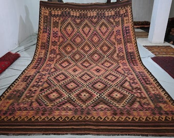 May include: A large rectangular woven rug featuring a repeating diamond pattern in shades of brown, rust orange, and muted pink. The design includes intricate geometric borders and a flat-weave texture, displaying a symmetrical arrangement of shapes.