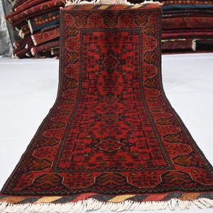 May include: A red and black patterned rug with a central medallion design. The rug has a rectangular shape with a detailed border and fringed edges. The rug is displayed on a white surface.