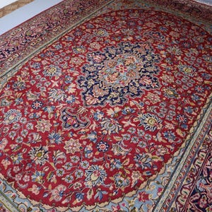 May include: A rectangular area rug featuring a deep red central field filled with intricate floral patterns in shades of blue, cream, and pink. The rug has a prominent dark blue central medallion and a wide, multi-layered border with ornate designs.