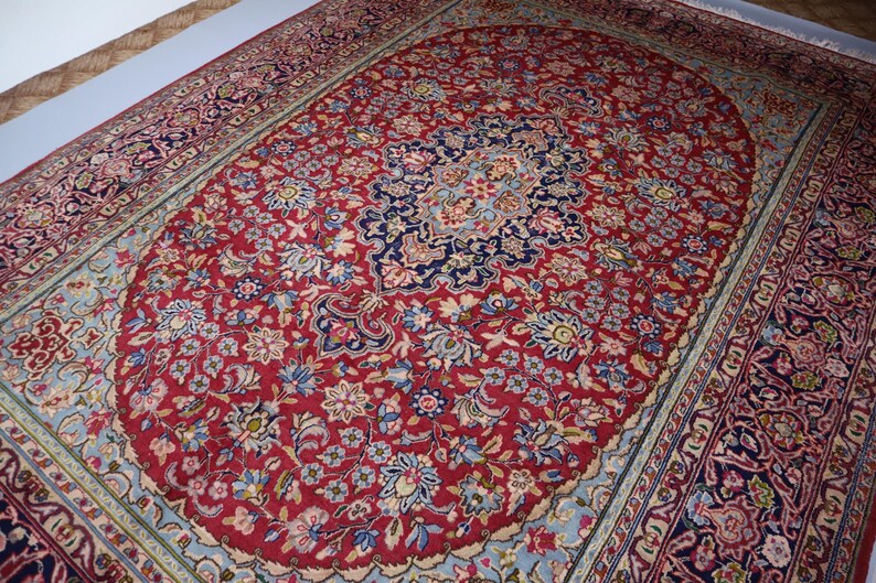 May include: A rectangular area rug featuring a deep red central field filled with intricate floral patterns in shades of blue, cream, and pink. The rug has a prominent dark blue central medallion and a wide, multi-layered border with ornate designs.