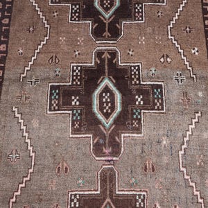 K&ouml;nnte beinhalten: Nahaufnahme eines braunen und beigefarbenen Teppichs mit einem zentralen geometrischen Muster. Der Teppich weist ein sich wiederholendes Design mit dunkelbraunen, wei&szlig;en und t&uuml;rkisfarbenen Akzenten auf. Die R&auml;nder haben eine dekorative Bord&uuml;re.