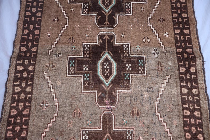 K&ouml;nnte beinhalten: Nahaufnahme eines braunen und beigefarbenen Teppichs mit einem zentralen geometrischen Muster. Der Teppich weist ein sich wiederholendes Design mit dunkelbraunen, wei&szlig;en und t&uuml;rkisfarbenen Akzenten auf. Die R&auml;nder haben eine dekorative Bord&uuml;re.