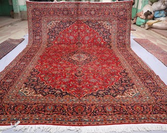 May include: A large, ornate red rug with a central medallion design and intricate floral patterns. The rug features a dark blue border and a cream-coloured fringe. Additional smaller rugs are visible.