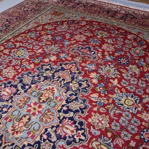 May include: A rectangular area rug featuring a deep red central field filled with intricate floral patterns in shades of blue, cream, and pink. The rug has a dark blue border with detailed botanical motifs and white fringe along the short edge.