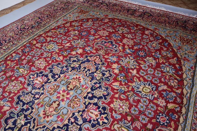 May include: A rectangular area rug featuring a deep red central field filled with intricate floral patterns in shades of blue, cream, and pink. The rug has a dark blue border with detailed botanical motifs and white fringe along the short edge.