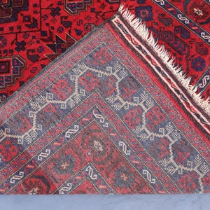 May include: A detailed shot of a red and blue patterned rug. The rug features intricate geometric designs in shades of red, blue, and cream. The rug's fringe is visible along one edge, and the rug is folded over to reveal the underside.