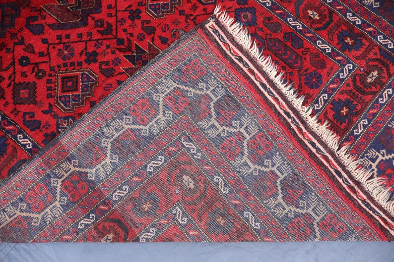 May include: A detailed shot of a red and blue patterned rug. The rug features intricate geometric designs in shades of red, blue, and cream. The rug's fringe is visible along one edge, and the rug is folded over to reveal the underside.