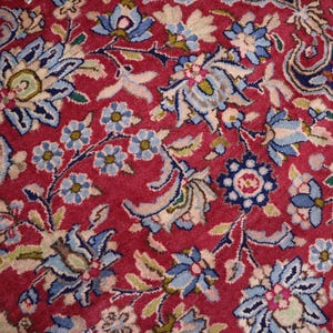 May include: A woven rug featuring a deep red background densely patterned with intricate floral motifs in shades of light blue, navy, cream, and sage green. The design consists of repeating blossoms, scrolling vines, and stylised leaves.