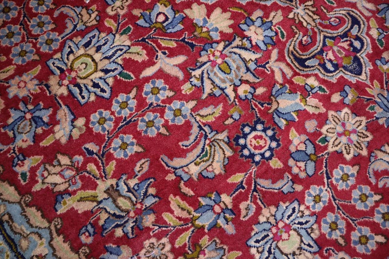 May include: A woven rug featuring a deep red background densely patterned with intricate floral motifs in shades of light blue, navy, cream, and sage green. The design consists of repeating blossoms, scrolling vines, and stylised leaves.