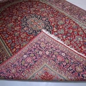 May include: A rectangular area rug featuring a deep red central field with a dark blue medallion and intricate floral patterns in shades of pink, light blue, and cream. One corner is folded over to reveal a contrasting underside with floral motifs.