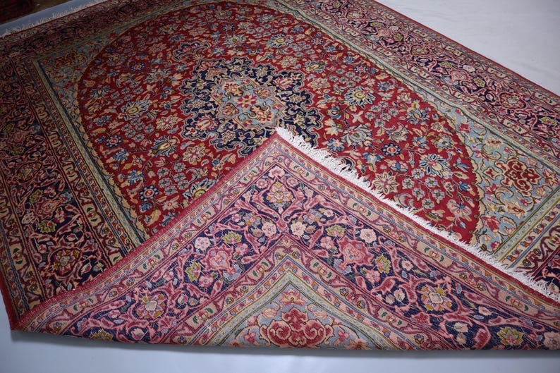 May include: A rectangular area rug featuring a deep red central field with a dark blue medallion and intricate floral patterns in shades of pink, light blue, and cream. One corner is folded over to reveal a contrasting underside with floral motifs.