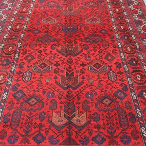 May include: A vibrant red rug with an intricate geometric pattern. The rug features a central design with repeating shapes in shades of brown and blue, framed by a detailed border. The rug's rich colors and complex design create a visually appealing textile.