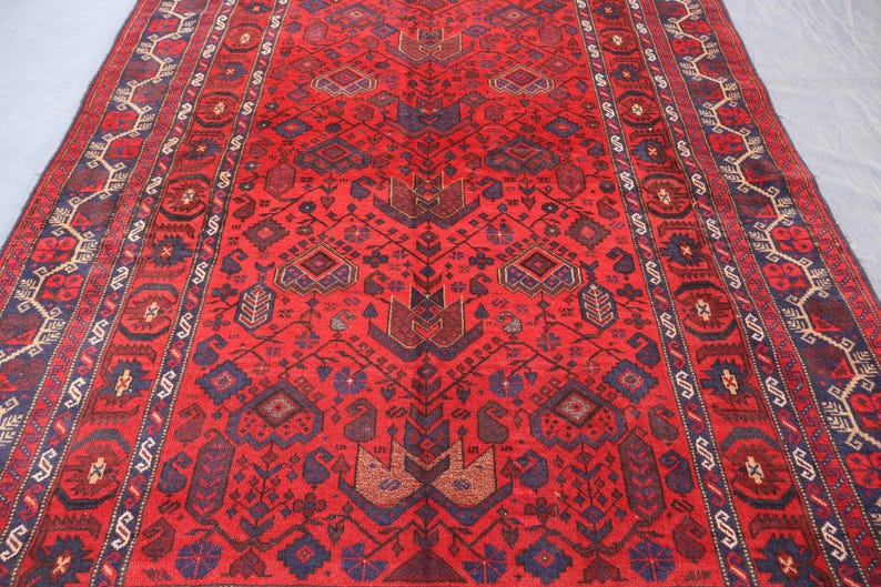 May include: A vibrant red rug with an intricate geometric pattern. The rug features a central design with repeating shapes in shades of brown and blue, framed by a detailed border. The rug's rich colors and complex design create a visually appealing textile.