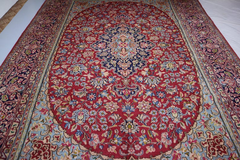 May include: A rectangular area rug featuring a deep red central field with an intricate navy blue and light blue floral medallion. The border displays a complex pattern of repeating floral motifs in shades of red, navy, and light blue.