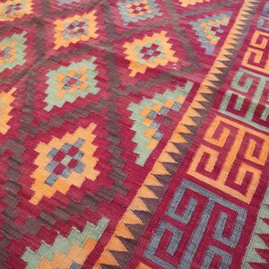 May include: A close-up of a red, orange, and green patterned rug. The rug features geometric designs in shades of red, orange, and green. The border has a repeating geometric pattern. The rug is likely made of woven fabric.