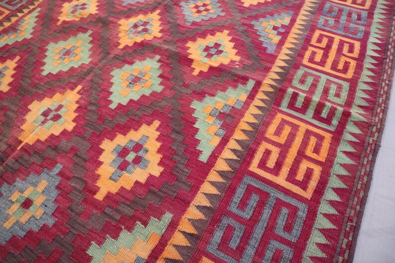May include: A close-up of a red, orange, and green patterned rug. The rug features geometric designs in shades of red, orange, and green. The border has a repeating geometric pattern. The rug is likely made of woven fabric.