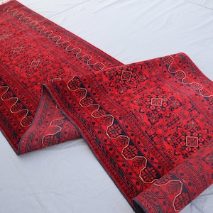 May include: A red and black patterned runner rug with a traditional design. The rug is rolled up and ready to be unfurled.