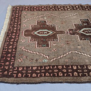 K&ouml;nnte beinhalten: Ein rechteckiger, brauner Teppich mit geometrischem Muster. Der Teppich hat ein zentrales Design mit sich wiederholenden Formen in Braun-, Beige- und T&uuml;rkist&ouml;nen. Der Rand ist dunklerbraun mit einem sich wiederholenden Muster. Der Teppich hat an einem Ende Fransen.
