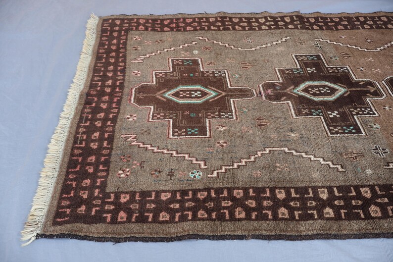 K&ouml;nnte beinhalten: Ein rechteckiger, brauner Teppich mit geometrischem Muster. Der Teppich hat ein zentrales Design mit sich wiederholenden Formen in Braun-, Beige- und T&uuml;rkist&ouml;nen. Der Rand ist dunklerbraun mit einem sich wiederholenden Muster. Der Teppich hat an einem Ende Fransen.