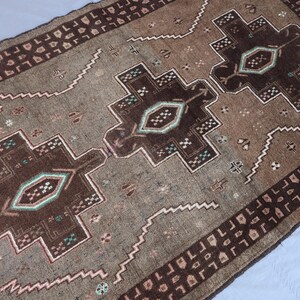 K&ouml;nnte beinhalten: Ein rechteckiger, handgewebter Teppich mit einem zentralen geometrischen Design in Braun-, Beige- und T&uuml;rkist&ouml;nen. Der Teppich hat ein sich wiederholendes Bord&uuml;renmuster und komplizierte Details. Die Abmessungen des Teppichs sind im Bild nicht sichtbar.