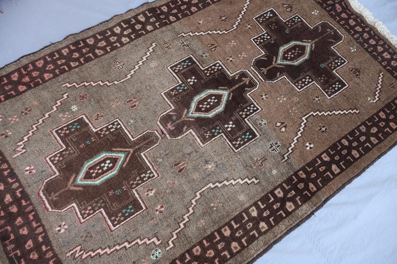 K&ouml;nnte beinhalten: Ein rechteckiger, handgewebter Teppich mit einem zentralen geometrischen Design in Braun-, Beige- und T&uuml;rkist&ouml;nen. Der Teppich hat ein sich wiederholendes Bord&uuml;renmuster und komplizierte Details. Die Abmessungen des Teppichs sind im Bild nicht sichtbar.