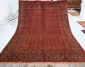 Könnte beinhalten: Ein großer rechteckiger Teppich mit einem tiefroten Feld und einem sich wiederholenden geometrischen Muster in Schwarz und Dunkelbraun. Der Teppich ist von einer breiten Bordüre mit komplexen floralen und geometrischen Motiven umrahmt und hat helle Fransen.